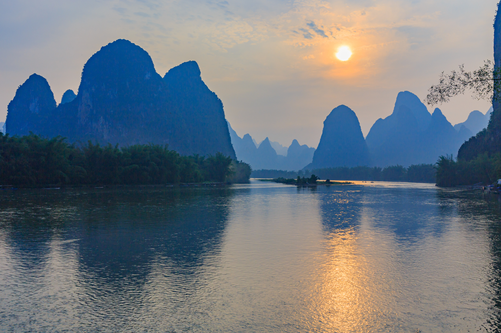 Sunset over a calm river with tall, jagged karst mountains along the shore, reflecting in the water