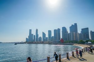 City skyline along a harbor with glass skyscrapers under a clear blue sky on a sunny day, viewed from the water's edge.
