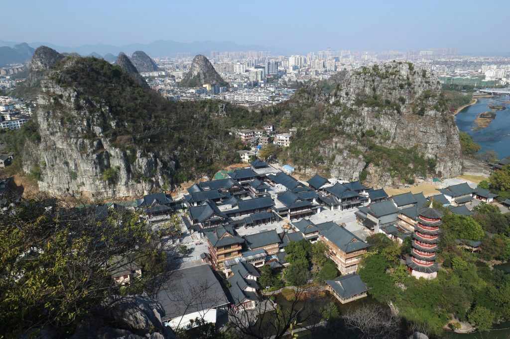 Limestone hills with sparse trees frame a cluster of traditional Chinese buildings at the base, with a city skyline in the distance and a river to the right.