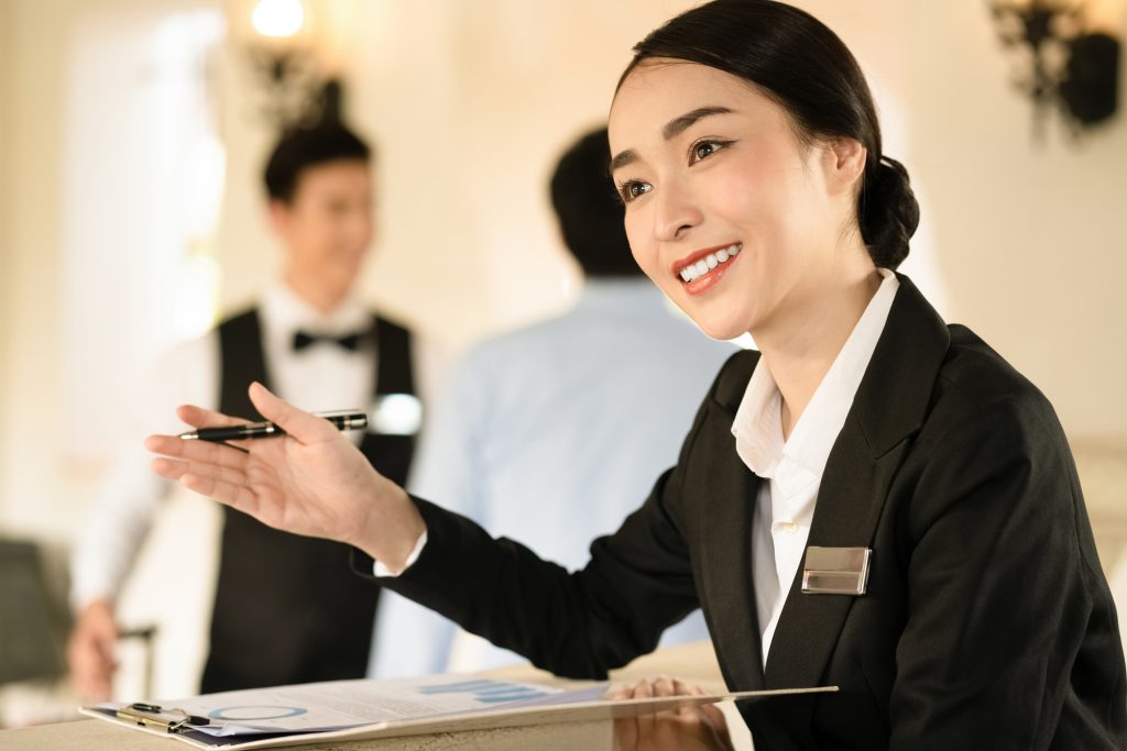 Smiling female receptionist in a black blazer extends a pen at a front desk toward a guest.