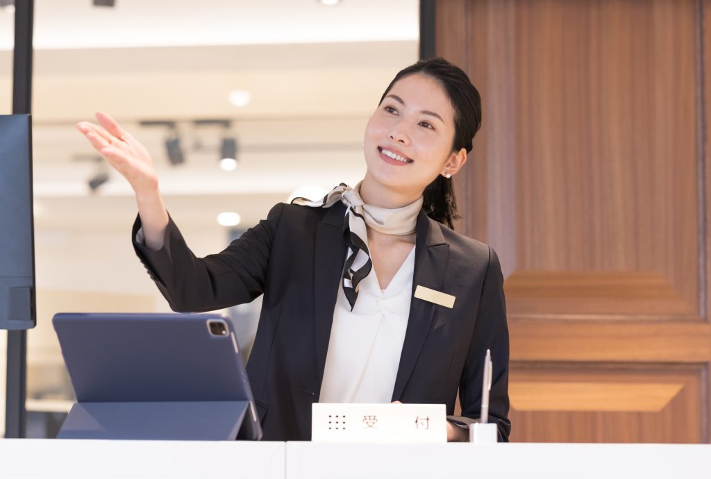 Receptionist in a dark blazer greets a guest at a front desk, with a tablet on the counter and a name badge visible.