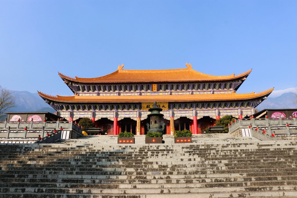 Temple with orange-tiled roof atop stone steps, red columns, and decorative eaves against a blue sky.